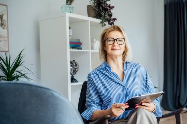 Profesyonel psikoterapi. Ofisteki koltukta oturan başarılı kadın psikolog kameraya gülümsüyor. 50 'li yaşlarda orta yaşlı bir öğretmen, koç, akıl hocası, terapist, danışman portresi.