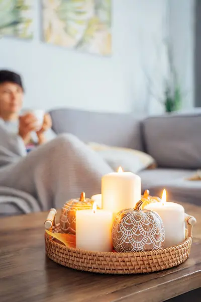 Focus on autumn fall cozy mood decor composition. Orange pumpkins, burning candles with a blurred woman covered with plaid and drinking tea on the sofa. Movie night party at home. Cozy relaxing time
