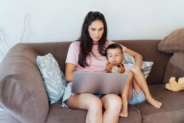 Young Mother and son on the sofa using laptop. Happy mom and little boy hugging and watching videos, reading books, playing video game, using online app, studying together at home. Family time