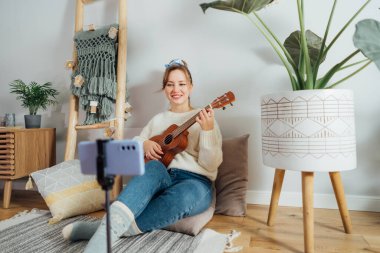 Genç kadın ukulele gitar çalıyor ve modern Scandi iç mekanında sosyal ağlar için telefonda Noel müzik videosu kaydediyor. Müzisyen blogcu şarkı söylüyor, içerik yaratıyor. Seçici odak