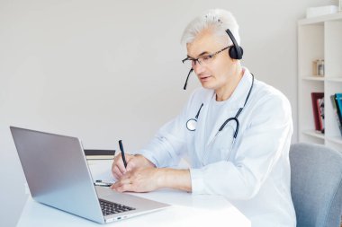 Orta yaşlı erkek doktor, hastalara internet üzerinden danışmanlık yapmak için kulaklık ve laptop kullanıyor. Ev sağlığı tedavisi için teletıp konsepti. Uzaktan kumandalı tıbbi randevu. Tıbbi teknoloji