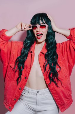Koyu yeşil, uzun saçlı, parlak kırmızı dudaklı, parlak bir moda kızı pembe arka planda üst ve güneş gözlüğü takıyor. Şık saç stili, makyaj. Seksi Model, Oyuncu Hippi Kadın, Yüzsüz Duygu