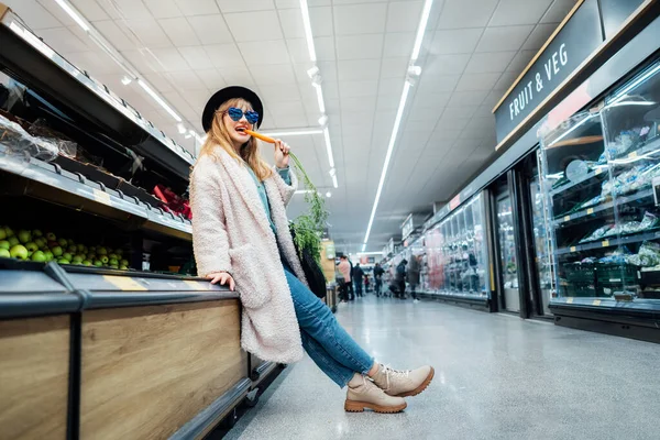 Stylish fashion smiling woman with fresh carrot in the supermarket store during selecting fresh products. Healthy eating diet, go vegan. Grocery Shopping. Veganuary diet month. Selective focus