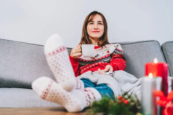 Relaxed woman in plaid and warm socks drinking hot tea with remote controller on sofa at home while watching movie, TV with christmas decoration atmosphere. Cozy and comfortable winter concept.