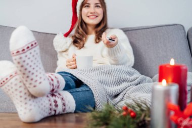 Uzaktan kumandalı, bulanık, Noel Baba şapkalı, sıcak çoraplı, rahat koltukta oturup film izleyen, Noel dekorasyonlu televizyon izleyen bir kadın. Sıcak kış.
