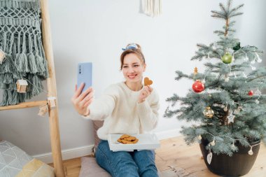 Modern Scandi 'nin evindeki saksıdaki yılbaşı ağacının yanındaki yerde dinlenirken rahat bir kazakla selfie çeken mutlu genç kadın. Sıcak kış tatili. İçerik yaratmak. Seçici odak