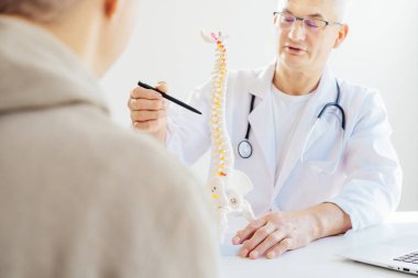 Orta yaşlı erkek doktorun işaret ettiği insan omurga modeline odaklanın ve hastanın klinikteki sorununu açıklayın. Omurga sağlığı ve sırt ağrısı tedavisi konsepti. Seçici odak, boşluğu kopyala.