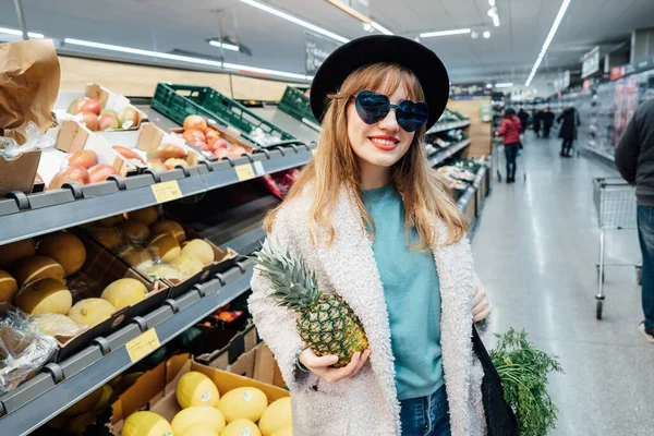 Stylish fashion smiling woman holding fresh organic pineapple in the supermarket store during selecting fresh products. Grocery Shopping. Healthy eating diet, go vegan. Selective focus. Copy space