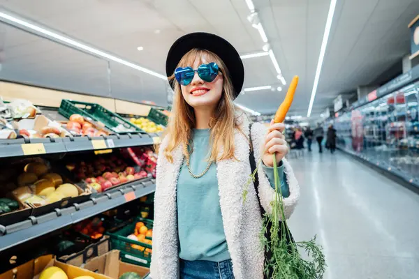 Stylish fashion smiling woman with fresh carrot in the supermarket store during selecting fresh products. Healthy eating diet, go vegan. Grocery Shopping. Veganuary diet month. Selective focus