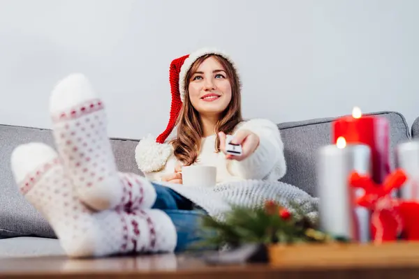 Relaxed, smiling woman in santa hat, jumper and warm socks sitting on comfortable couch, using tv remote controller and watching movie, TV with christmas decoration atmosphere. Cozy winter concept