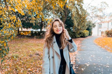 Trençkotlu, gözleri kapalı, gülümseyen romantik genç bir kadın ara verir. Sonbahar parkında sarı yapraklarla yürüyüşün tadını çıkarıyordu. Romantik sonbahar havası. Boşluğu kopyala.