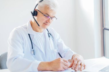 Orta yaşlı erkek doktor, hastalara internet üzerinden danışmanlık yapmak için kulaklık ve laptop kullanıyor. Ev sağlığı tedavisi için teletıp konsepti. Uzaktan kumandalı tıbbi randevu. Tıbbi teknoloji