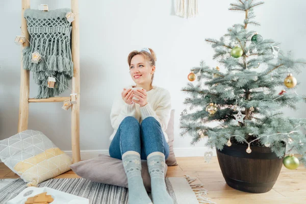 Happy Young Woman in cozy sweater with cup of hot drink relaxing on the floor cushions near potted christmas tree in modern Scandi interior home. Eco-friendly cozy winter holidays. Selective focus
