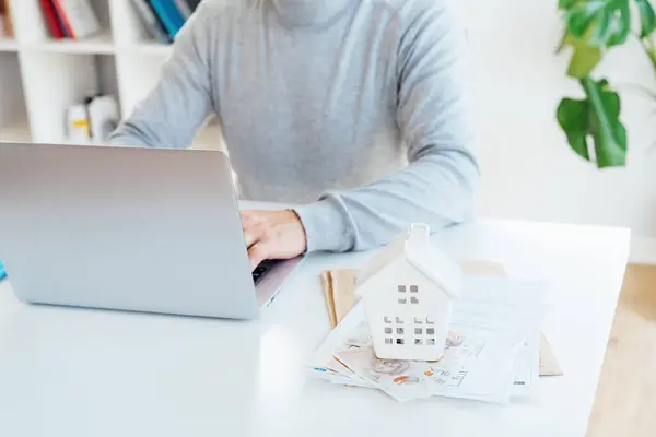 Focus on House model, money and docs on table and blurred senior man works on laptop. Housing cost and payments. Real estate, property investment. Home mortgage loan rate. Saving money for retirement.