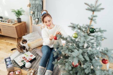Kedisi aydınlık İskandinav iç mekanında onu izlerken rahat süveter süveter süsleme saksısı Noel ağacı içindeki genç kadın. Çevre dostu kış tatili. Evcil hayvan evde. Seçici odak