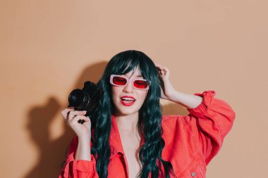 Koyu yeşil, uzun saçlı, parlak kırmızı dudaklı, gözlüklü, bej arka planda retro film kamerası tutan küstah bir moda kızı. Moda genç hipster fotoğrafçı, seyahat fotoğrafı.