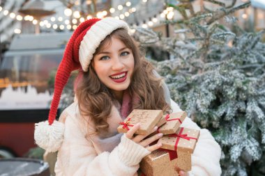 Noel Baba şapkalı genç bir kadın geleneksel şehir caddesi Noel pazarında hediye kutularıyla dolu bir kuleye sahip. Mutlu kadın Noel hediyelerini seçiyor. Mutlu Noeller. Mutlu yıllar. Alışveriş, satış, indirim.
