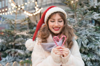 Noel Baba şapkalı genç bir kadın geleneksel Noel pazarında yürürken baston şekerlerle kalp şeklinde kalp yapıyor. Mutlu yıllar Noel tatilleri. Aşkı paylaşın, hayırseverlik konsepti.