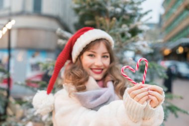 Geleneksel Noel pazarında Noel Baba şapkalı genç bir kadının baston şekerlerinden yapılmış kalp şekline odaklan. Mutlu yıllar Noel tatilleri. Aşkı paylaşın, hayırseverlik konsepti