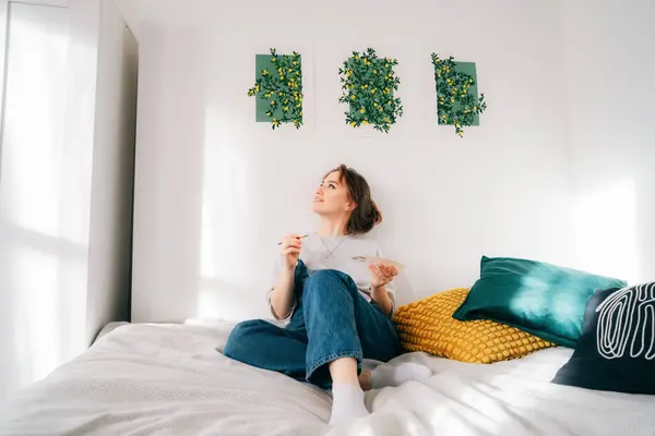 Smiling female artist sitting on bed with paintbrush, palette under her hand-drawn painting art works on the wall at home. Art, hobby, leisure and creative occupation concept. DIY, Home decorating