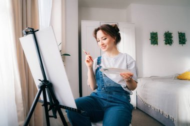 Kadın sanatçı sehpanın önünde oturuyor, esinlenerek resim çiziyor, boya fırçası, palet, gülümsüyor, evde dinleniyor. Sanat, hobi, eğlence ve yaratıcı meslek kavramı.