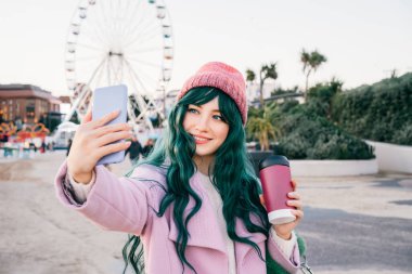 Tarz sahibi, genç, gülümseyen hippi bir kadın elinde tekrar kullanılabilir kahve fincanı tutan pembe bir ceket giyen, örgü şapkalı, telefonda telefonla fotoğraf çeken, mutlu ruh hali, mevsimsel moda eğilimi olan.