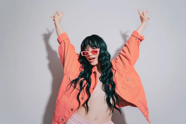 Cheeky emotional fashion young woman with long green color hair and trendy peach color lips, jacket and sunglasses dances on white background. Playful Hipster woman having fun. Color of the 2024 year