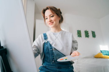 Kadın sanatçı sehpanın önünde oturuyor, esinlenerek resim çiziyor, boya fırçası, palet, gülümsüyor, evde dinleniyor. Sanat, hobi, eğlence ve yaratıcı meslek kavramı..
