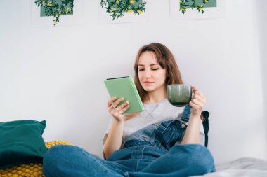 Yatakta tablet okuyucu hakkında e-kitap okuyan genç bir kadın. Kadın rahat bir odada elektronik kitap okurken sıcak içecekle rahatlıyor. Zihinsel sağlık, kişisel gelişim. Hobi, yalnız zaman geçirmek.