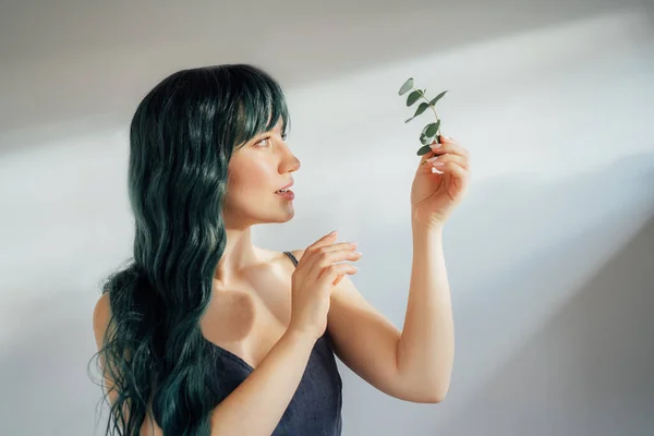 Portrait of Adorable tender young woman with long green color hair holding eucalyptus branch in hand in ray of sun from window on light background. Concept of beauty, art, fashion. Mermaid style