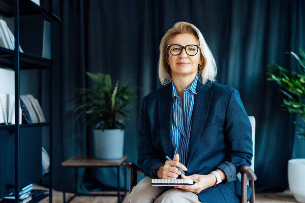 Портрет улыбающейся 50-х стильной, уверенной в себе зрелой предпринимательницы, директора компании среднего возраста, опытной старшей женщины-профессионала, руководителя команды бизнес-тренеров в современном офисе. Женщина-лидер.