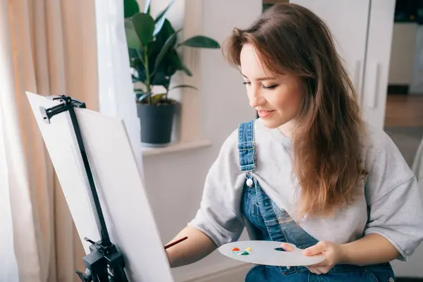 female artist sitting in front of easel, painting picture with inspiration, drawing on canvas with paintbrush, palette, smiling, relaxing at home. Art, hobby, leisure and creative occupation concept
