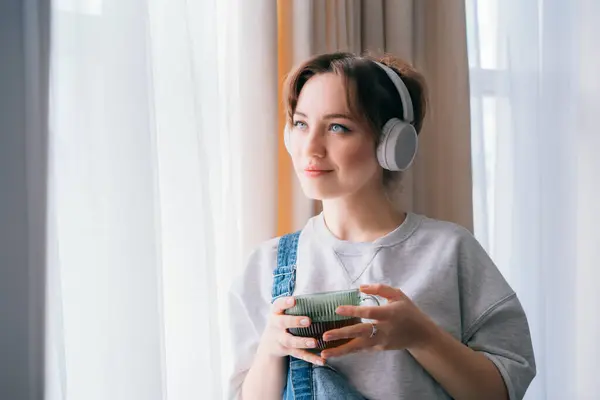 Young woman listen music, enjoy cup of coffee or tea and looks out window. Calm female spend free time at home enjoy favourite song with wireless headphones. Pastime weekend relax, no stress concept