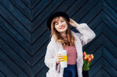 Portrait of young smiling fashion woman with reusable coffee cup and net bag with fresh tulip flowers inside on the black wooden background. Urban city street fashion. Spring mood. Selective focus.