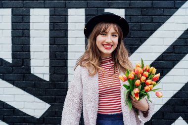 Mutluluğun portresi. Gülümseyen genç moda kadını duvarın arka planında bir buket taze lale çiçeği tutuyor. Şehir sokak modası. Bahar pozitif havası. Seçici odak.