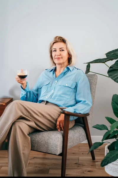 Portrait of confident elegant middle aged woman sitting in retro style armchair with cup, enjoying coffee. Smiling stylish senior 50s lady in modern living room with green plants. old money aesthetic.