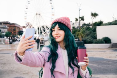 Tarz sahibi, genç, gülümseyen hippi bir kadın elinde tekrar kullanılabilir kahve fincanı tutan pembe bir ceket giyen, örgü şapkalı, telefonda telefonla fotoğraf çeken, mutlu ruh hali, mevsimsel moda eğilimi olan.