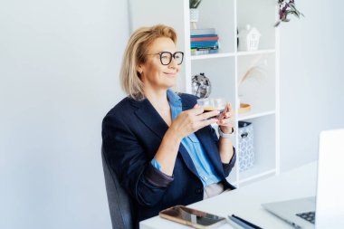 Başarılı, rahat, rüya gibi orta yaşlı iş kadını iş yerinde dinleniyor, kahve molasının tadını çıkarıyor. Gülümseyen, kendinden emin orta yaşlı tecrübeli, son sınıf bayan profesyonel kahvesini içiyor..