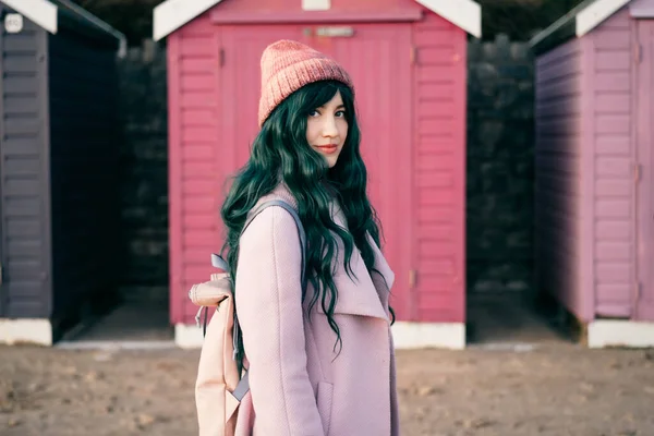 Stylish hipster woman with color hair in pink outfit and backpack walking along wooden beach huts on seaside. Off season Travel concept. Seasonal street fashion. Barbiecore style. Simple pleasures