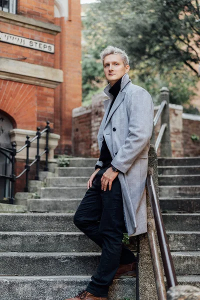 Handsome male model with gray hair in a black roll neck jacket and trousers, checked coat walking on the old city street. Cold season fashion stylish outfit trend. Old money aesthetic. Soft focus