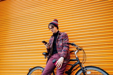 Genç hippi turuncu duvarın yanında kruvazör bisikleti üzerinde otururken cep telefonu kullanıyor. Öğrenci 5G ağı kullanıyor, bindikten sonra dinleniyor. Karbon ayak izi için çevre dostu taşımacılık. Sürdürülebilir yaşam tarzı.