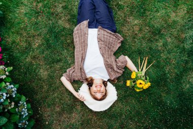 Üst manzara mutlu genç kadın bahçıvan yeşil çimlere uzanmış, sadece sarı ayçiçekleri toplamış, yukarı bakıyor ve gülümsüyor. Kadınlar anın tadını çıkarırlar, dışarıda dinlenirler. Bahar ya da yaz mevsimi konsepti.