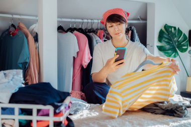 Elbiselerini satmak ya da bağışlamak için akıllı telefonunu kullanan bir kadın gömleğin fotoğrafını çekiyor. Temizlik, giysi ayıklama ve gardırop temizleme. Tekrar kullan, ikinci el konsept. Bilinçli tüketici.