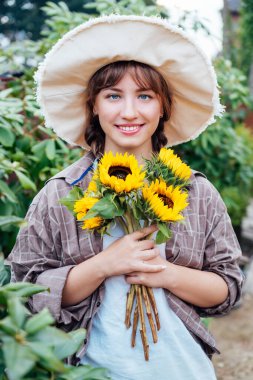 Yeşil ağaçların arkasında elinde taze ayçiçeği buketi tutan gülümseyen genç kadın çiftçi portresi. Kırsal, temel kır evi yaşam tarzı. Seçici odaklanma. Dikey kart.