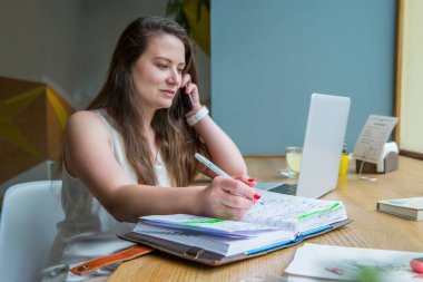 Cep telefonuyla konuşurken not defterine not alan ve kafede dizüstü bilgisayarla çalışan iş kadınına odaklan. Uzak bir işyeri. Freelancer çalışma alanı. Uzayı kopyala Seçici odak
