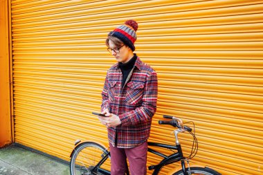 Genç hippi turuncu duvarın yanında bisiklet sürerken cep telefonu kullanıyor. Öğrenci 5G ağı kullanıyor, bindikten sonra dinleniyor. Karbon ayak izi için çevre dostu taşımacılık. Sürdürülebilir yaşam tarzı.