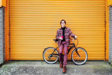 Genç, gülümseyen hippi erkek öğrenci turuncu duvarın yanında kruvazör bisikletinin üzerinde otururken termostan kahve içiyor. Karbon ayak izi için çevre dostu taşımacılık. Sürdürülebilir yaşam tarzı