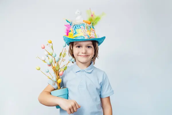 Easter eggs hunt tradition. Smiling boy kid in Easter hat holds easter tree in pot and chocolate egg in other hand on the white background. Easter decoration, family celebration, Christian traditions