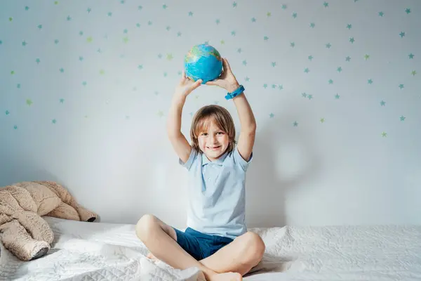 Smiling caucasian boy holding an earth globe model over his head sitting on the bed with stars background on the white wall. Save the planet, Earth Day. Future Global peace and unity concept.
