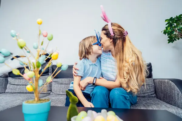 Happy smiling Caucasian mother and her son in bunny ears having fun at home on Easter day. Family sitting on sofa at home hugging, mother kissing son. Family Easter traditions. Selective focus.
